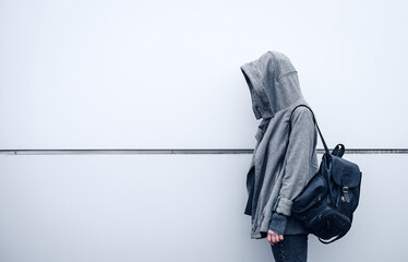 Girl without a face in a hood with long dark hair and a gray sweatshirt with a black leather backpack near a gray wall