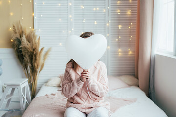 A woman in a pastel pink sweater sitting on the bed in the bedroom hid her face behind an...