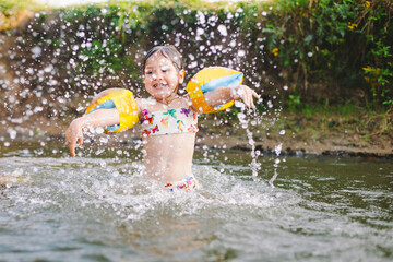 Happy summertime, healthy childhood concept. Little girl with yellow cushions splashing and having fun in a river in summer. Blurred green background. Horizontal shot.