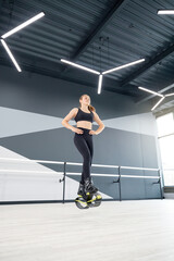 Fototapeta na wymiar From below view of girl practicing dance moves while doing kangoo jumps, hands on waist. Young female athlete with ponytail wearing tight black sportswear, cardio exercise in hall, hi tech interior.