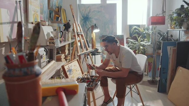 Tracking shot of young Caucasian man sitting in art studio and sharpening pencil with knife while preparing tools for drawing