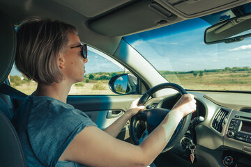 Fototapeta na wymiar Portrait of serious woman in sunglasses driving car in motion through fields in countryside