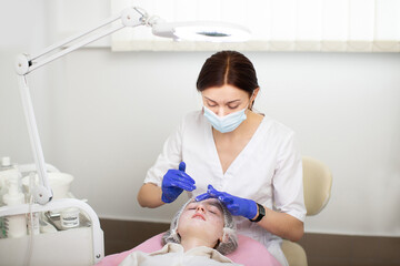 Female cosmetologist wearing face mask and medical gloves, applying cream or foam on a girl's face to clean her skin. A cosmetology procedure in a beauty salon for skin acne treatment