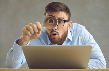 Portrait of funny handsome nerdy young business man in glasses sitting at office desk with laptop computer, holding magnifying glass and looking at something with big eye and surprised face expression - Powered by Adobe