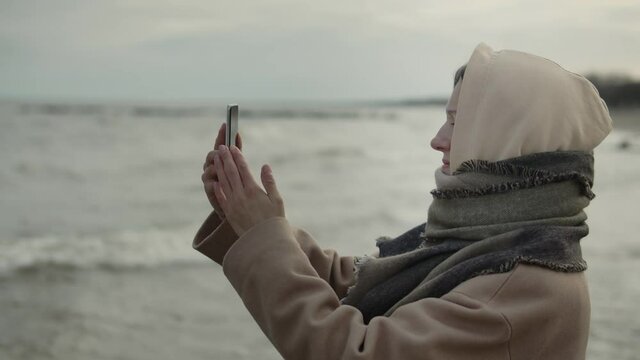 Woman wearing warm clothes takes photos with her smartphone on the beach by the sea. Girl in warm coat with hood on her head walking on empty beach