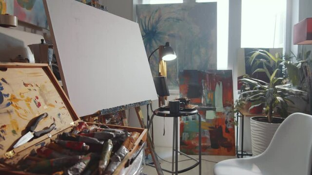 Zoom out shot of canvas on easel, chair and wooden case with painting tools in creative art studio with no people