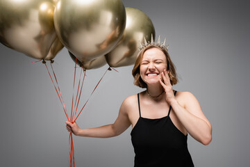 happy plus size woman in black slip dress and crown holding golden balloons on grey © LIGHTFIELD STUDIOS