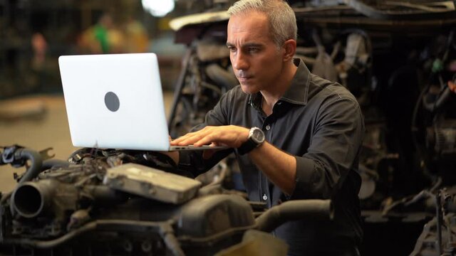 senior engineer using laptop computer checking list old used car engine in machinery parts shop warehouse. old technician man Repair vehicle automobile machine in Recycle motor factory