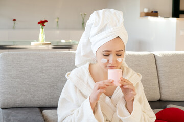 Young woman in white towel chilling on sofa and making nourishing mask or creme. Beauty treatment and spa relaxing at home. Morning skin care beauty routine, self care.