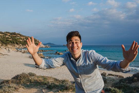 Portrait of happy joyful young latin man smiling looking at the camera with open arms with beautiful beach on background. Travel destination in Sardinia, Italy.
