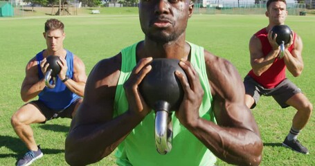 Diverse group of three fit men exercising outdoors, squat lifting kettlebell weights