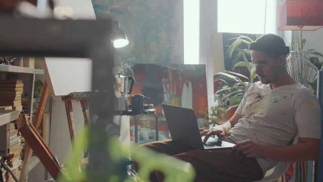 Young Caucasian man in stained t-shirt browsing the web on laptop and drinking tea in creative art studio