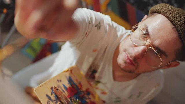Close up top down shot of handsome male painter in beanie hat, stained t-shirt and glasses painting picture with palette and brush