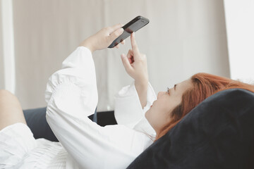 Woman in white dress lying on a sofa in the living room use smartphone. Concept of life and relax at home.	