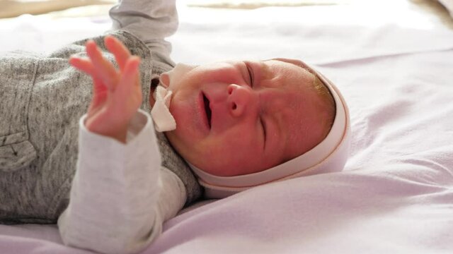 Very small baby girl lie on back at large bed alone and cry, shake hands shake hands. Clip with original newborn voice sound. Infant call for mother or feel stomach ache