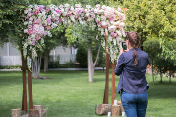 Girl taaking photo of very beautiful and stylish wedding arch, decorated with various fresh flowers, standing in the garden.