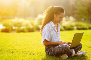 Young woman freelancer sitting on green grass with laptop. Education online. Happy woman with wireless headphones calling on laptop, talk by webcam, video conference.