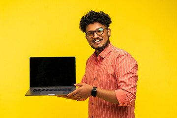 Attractive smiling Indian isolated on yellow background holding laptop with black screen and showing it to camera, yellow background, studio photo