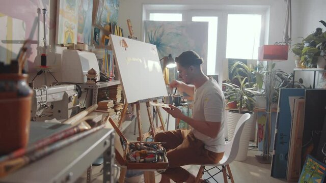 Zoom in shot of professional male artist holding wooden palette and applying paint to canvas with palette knife while working on new picture in studio