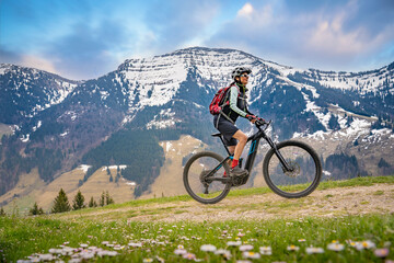 Fototapeta na wymiar pretty mid age woman riding her electric mountain bike in early springtime in the Allgau mountains near Oberstaufen,below the spectacular snow capped mount HochgratNagelfluh chain
