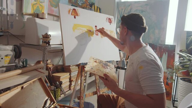 Zoom in shot of creative male artist holding wooden palette and applying paint to canvas with palette knife while painting new picture in studio