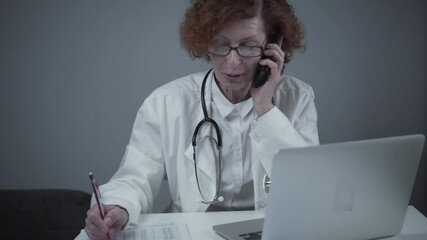 Caucasian senior female doctor physician talking on mobile at work. Healthcare professional answering call giving remote consultation on smart phone make appointment notes on laptop in office - Powered by Adobe