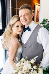 Handsome caucasian groom in a vest hugs european bride in white dress tender standing near window...