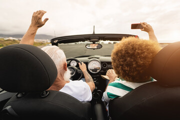 Trendy senior couple having fun taking selfie with smartphone inside convertible car - Multiracial...