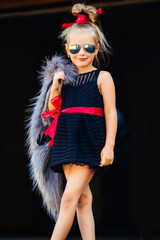 A spectacular little stylish girl in glasses and a black dress holds a fur vest with her handstands in the doorway.