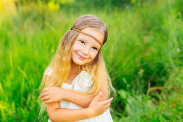 a charming, sweet, little girl with beautiful brown eyes, in a white dress, smiling, dressed in a hippie style, hugging her shoulders, against a background of green grass.