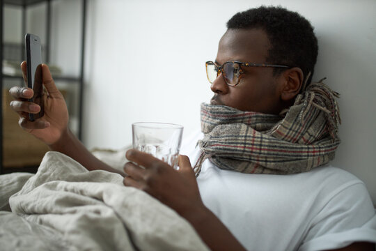 Sick young black man staying home because of respiratory disease texting message to boss on cell phone, taking medication with water, lying in bed wearing warm scarf around neck. Health and technology