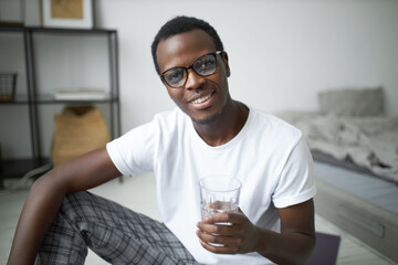Joyful handsome young black man in stylish glasses relaxing on floor after morning cardio workout,...