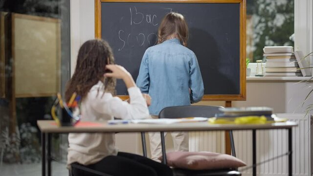 Happy schoolgirl writing Back to school on chalkboard in classroom with African American classmate sitting at desk. Cheerful schoolchildren rejoicing education reopening. Happiness and studying