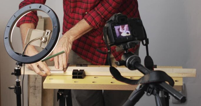 Vlogging, DIY guy drawing marks on wood plank using square ruler, and start cutting it using hand saw, recording it using smartphone with ring light and digital slr camera, real people, close-up