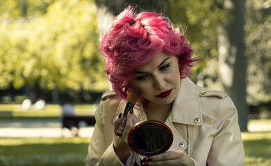 Mujer maquillándose en el parque , mujer de pelo rosa mirándose en el espejo, retocándose el maquillaje 