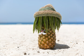 Ripe and juicy mustachioed pineapple in a hippie hat stands on the beach on white sand, in the...