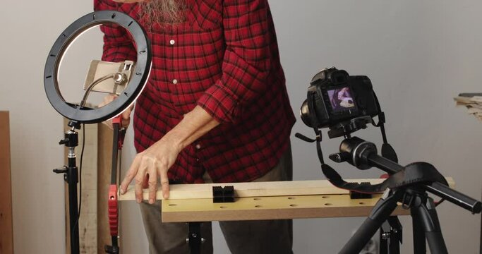 Vlogging, DIY guy cutting wood plank using hand saw, recording it using smartphone with ring light and digital slr camera