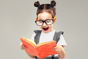 Child girl reads a book in surprise. A kid in glasses looks in amazement at an orange notebook....