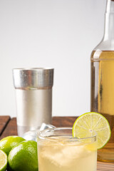 Brazilian drink caipirinha, glass of caipirinha, bottle of cachaça, cocktail shaker and lemons on wooden mat and rustic wood light background, selective focus.