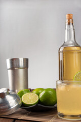 Brazilian drink caipirinha, glass of caipirinha, bottle of cachaça, cocktail shaker and lemons on wooden mat and rustic wood light background, selective focus.
