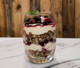 Nutrition and sweet desserts. Closeup view of a multilayer trifle with berries, oatmeal, granola, blueberries, raspberries and cream, served in a glass cup on the white marble table.