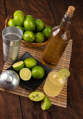 Brazilian drink caipirinha, glass of caipirinha, bottle of cachaça, cocktail shaker and lemons on wooden mat and rustic wood light background, selective focus.