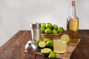 Brazilian drink caipirinha, glass of caipirinha, bottle of cachaça, cocktail shaker and lemons on wooden mat and rustic wood light background, selective focus.
