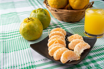 Orange ponkan, orange ponkan slices on a black plate, plus some oranges and a glass of juice on a checkered tablecloth on a light background, selective focus.