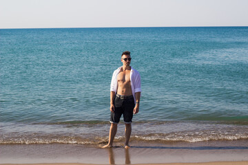 Ein hübscher, jünger Mann steht am Ufer des Oceans in der Türkei. Seit Hemd ist geöffnet, man sieht sein durchtrainierten Körper, Er hat Sonnenbrille auf und kurze Hose. Er schaut zur Seite. 