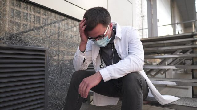 Worried and tired doctor sitting on stairs outside hospital. Exhausted physician in white lab coat and medical protective mask, sit on staircase clinic. Stress healthcare worker during COVID-19