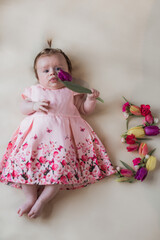 Cute little toddler girl in a pink dress. Lying on a bed with tulip flowers. Looking at the camera. Floral number.