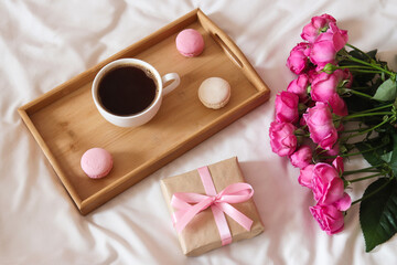 Fototapeta na wymiar Cozy holiday morning composition with roses, gift box and cup of coffee with macaroons on wooden tray. Mother's Day concept