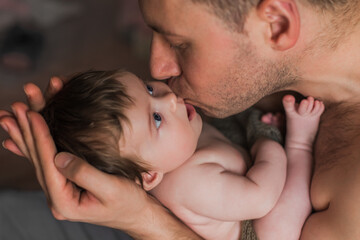 Naked father holds a naked tiny baby in his arms kissing him. 3 months. Indoor.
