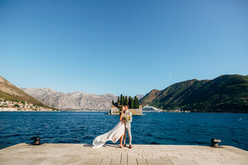 The bride and groom are embracing on the pier in the Bay of Kotor, behind them are mountains, island and old town of Perast